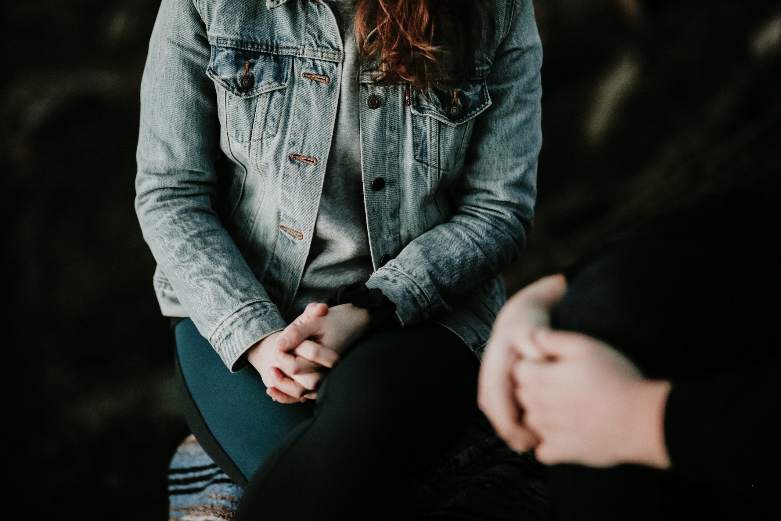 person sitting down wearing a denim jacket, with their hands on their lap, you can't see their face. There's another person in the foreground, we can only see their hands also on their lap