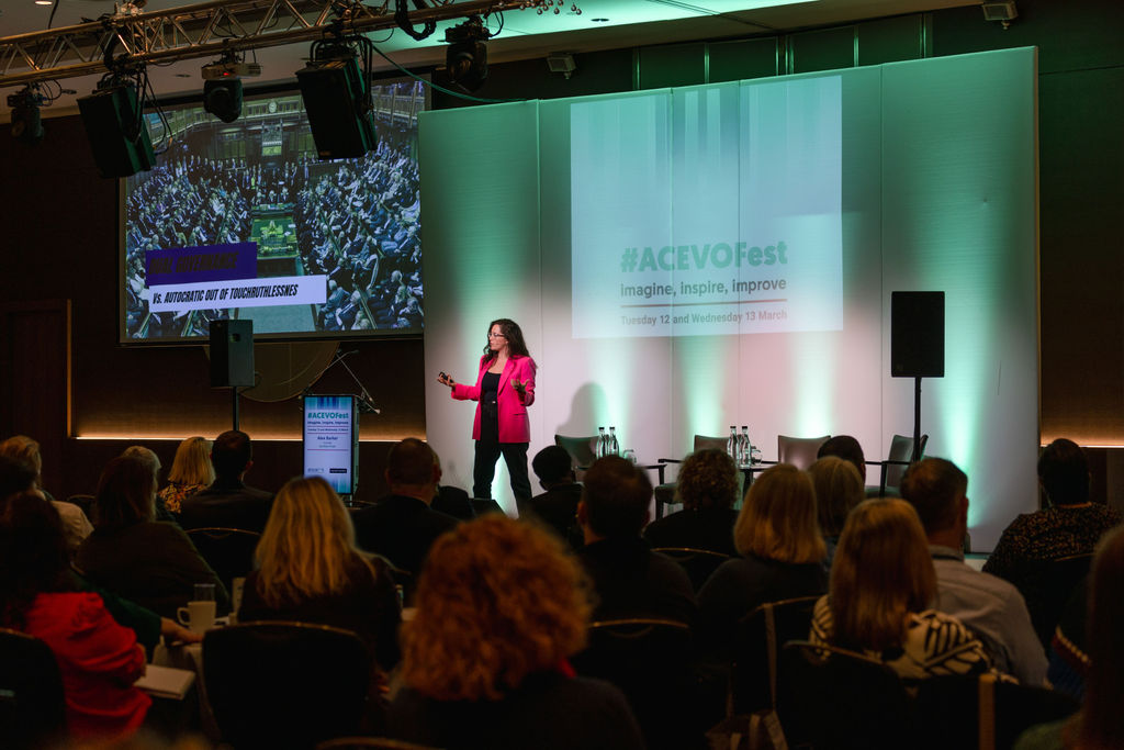 A photo of a woman stood on a stage speaking to a room of people