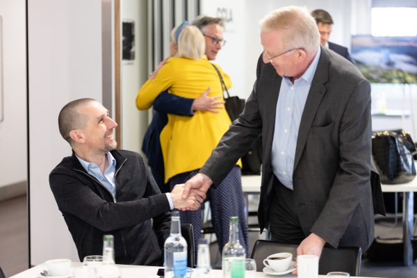 A man is sat at a table and is shaking hands with a man standing up, two people hug in the background