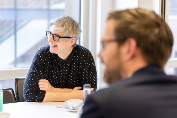 A woman is sat at a table, she is looking to the right and smiling