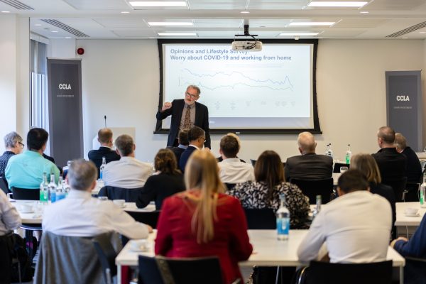 A man standing up, presenting at the front of a room, a group of people are sat listening